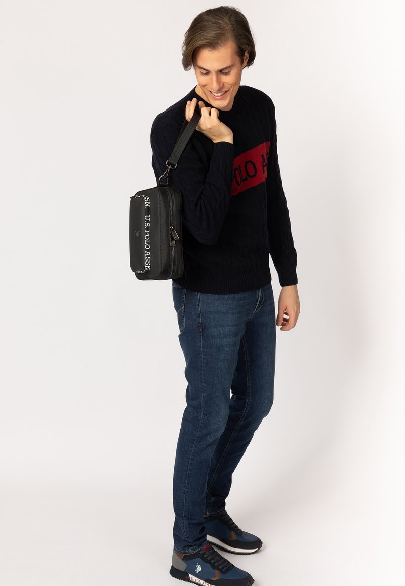 Sort crossbody taske med en glat tekstur, der pryder logoet "US Polo Assn." i hvidt, parret med en marineblå kabelstrikket sweater og jeans.