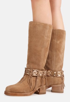 Taupekleurige suède kniehoge laarzen met decoratieve gevlochten riemen en metalen munten, gedragen op benen met gestapelde houten hakken.