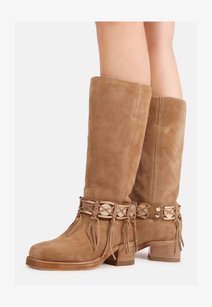 Botas altas hasta la rodilla de ante color canela con correas trenzadas decorativas y monedas de metal, usadas en piernas con tacones de madera apilados.