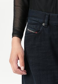 Χέρι χαλαρό κοντά στο ισχίο, φορώντας διαφανή μαύρη μπλούζα με μακριά μανίκια και σκούρα τζιν με ορατή ετικέτα Diesel Industry στην μπροστινή τσέπη.