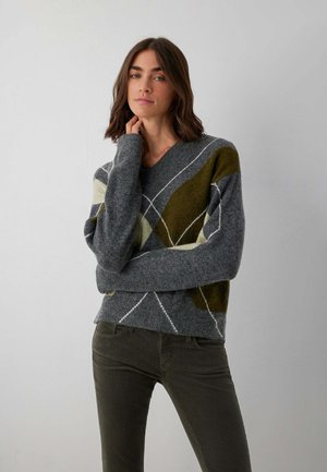 Frau mit schulterlangem braunem Haar trägt einen grauen Pullover mit weißen und olivgrünen geometrischen Mustern und dunkelgrünen Hosen, steht vor einer schlichten Wand.