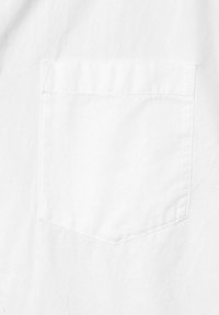 Biała bawełniana tkanina o delikatnej fakturze ma jedną kwadratową kieszonkę z przeszytymi brzegami po lewej stronie.