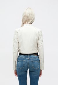 Valge nahast mootorratturi jakk, millel on tikitud õlavarre detail, lühendatud disain ja lukud varrukaotsades, koos siniste teksadega.