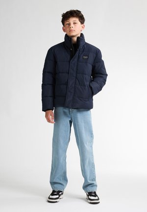 Veste matelassée marine avec un col haut, présentant un design matelassé et une fermeture éclair à l'avant. Associée à un jean bleu clair et des baskets noires.