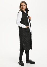 Gilet matelassé noir avec un col haut, des poches latérales et des accents à boutons-pression. Porté sur une chemise blanche et un pantalon noir, avec une coupe longue.