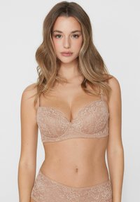 Žena s dlouhými vlnitými vlasy, která nosí bežový květinový krajkový set podprsenky a kalhotek, stojí před jednobarevným světlým pozadím.