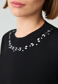 Schwarzer Strickpullover mit Rundhalsausschnitt, verziert mit aufwendigen Verzierungen aus klaren Edelsteinen und schwarzen Akzenten um den Kragen.