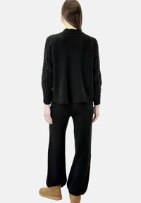 Suéter negro de punto con mangas de trenzado, combinado con pantalones de pierna ancha negros y calzado marrón claro. Textura suave, diseño relajado.