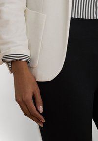 Blazer bianco in tessuto strutturato, con una tasca e dettagli a manica a righe. Abbinato a pantaloni neri aderenti e una camicia a righe sotto.