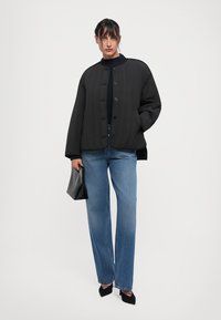 Frau trägt eine schwarze knöpfbare Jacke, einen dunklen Pullover, blaue weit geschnittene Jeans, schwarze Absätze und hält mit einer Hand eine schwarze Handtasche, während die andere Hand in der Jackentasche ist.