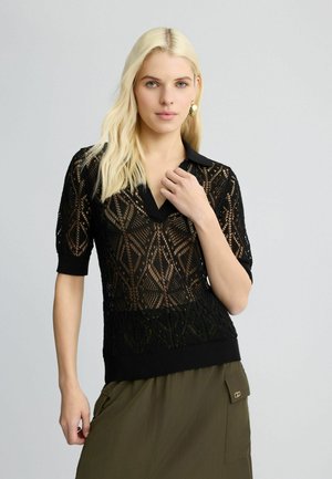 Polo en tricot noir avec des manches courtes, présentant un motif géométrique ajouré et un ourlet côtelé. Assorti à une jupe olive.