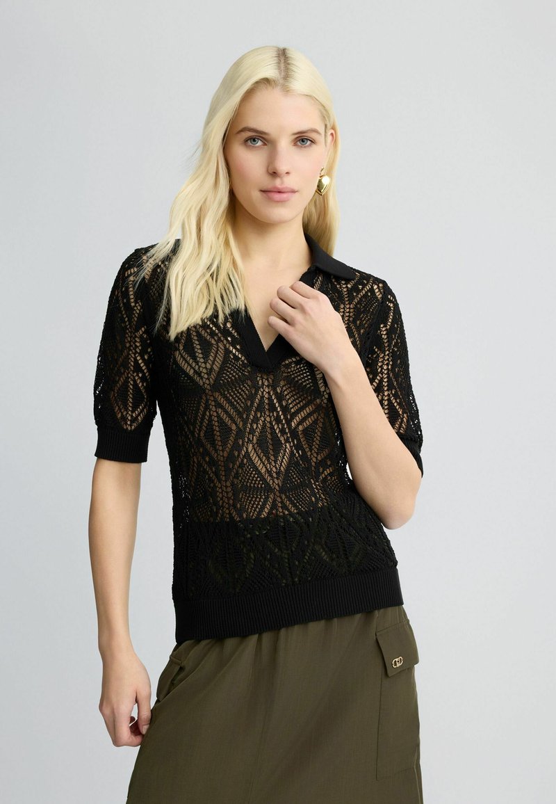 Maglione polo lavorato a maglia nera con maniche corte, caratterizzato da un motivo geometrico a rete e un orlo a costine. Abbinato a una gonna oliva.