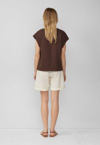 Woman with long blonde hair wearing a brown sleeveless top, white rolled-up shorts, and brown sandals, standing against a plain gray background.