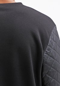 Maglione nero con maniche in pelle trapuntata visto da dietro, che mette in evidenza la texture del tessuto e i dettagli delle cuciture delle maniche.