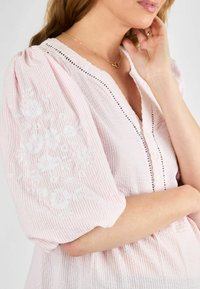 Rožinė dryžuota palaidinė su pūstomis rankovėmis, į kairę rankovę ploniai siuvinėtomis baltomis gėlėmis, V formos iškirpte ir mygtukų uždarymu priekyje.