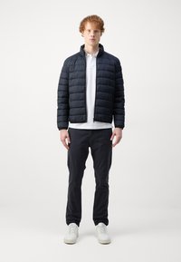 Navy puff jacket with quilted texture, featuring a stand-up collar, zip closure, paired with a white polo and dark pants. White sneakers complete the look.