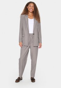 Světle šedý šitý oblek obsahující blazer se dvěma předními kapsami a odpovídajícími elastickými kalhotami. Nositelný přes bílý top a v kombinaci s hnědými botami.
