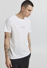 Hvid kortærmet t-shirt lavet af bomuld med en rund halsudskæring. Har et trykt logo, "reflect," i en kontrastfarve foran.