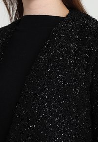 Černý kabát s texturou a jemným, třpytivým povrchem. Má volný střih a otevřený přední díl, vrstvený přes jednoduché černé tričko.