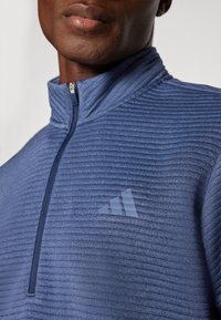 Pullover in fleece blu con zip, dotato di una texture a righe orizzontali, colletto alto e un delicato logo Adidas sul petto in una tonalità più chiara.