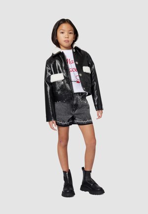 Veste en cuir noire cropped avec des accents en fourrure blanche, assortie à un short en denim gris et des bottines noires à zip à la cheville. T-shirt graphique blanc en dessous.