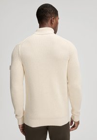Sweter w kolorze kremowym, wykonany na drutach, z wysokim kołnierzem, teksturowanym wzorem oraz ściągaczami na mankietach i dolnym brzegu. Posiada dopasowany krój i nieco wydłużony tył.