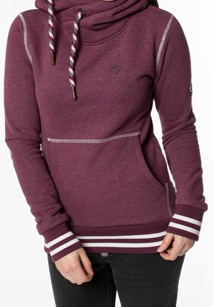 Femme portant un sweat à capuche bordeaux avec des poignets rayés, un emblème en forme de cœur sur la poitrine et des cordons rayés, associé à un pantalon noir.