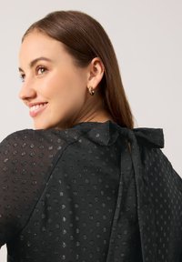 Schwarze Bluse mit strukturiertem Pünktchenmuster, durchscheinendem Stoff und einer Schleifenakzent am rückseitigen Halsausschnitt. Verfügt über kurze Ärmel und eine glatte Oberfläche.