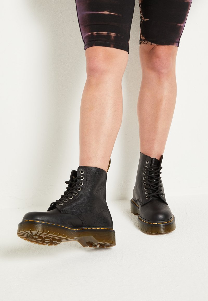 Personne portant des bottes montantes en cuir noir à lacets avec des semelles épaisses translucides et un short noir à motifs, debout sur un sol blanc.