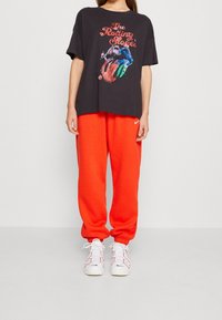 Black graphic t-shirt with The Rolling Stones logo, paired with orange sweatpants and white athletic shoes with laces and logo accents.