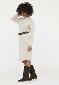 Robe tricotée beige avec des manches longues, des poignets côtelés et des accents de boutons à l'épaule, assortie à des bottines en daim marron.