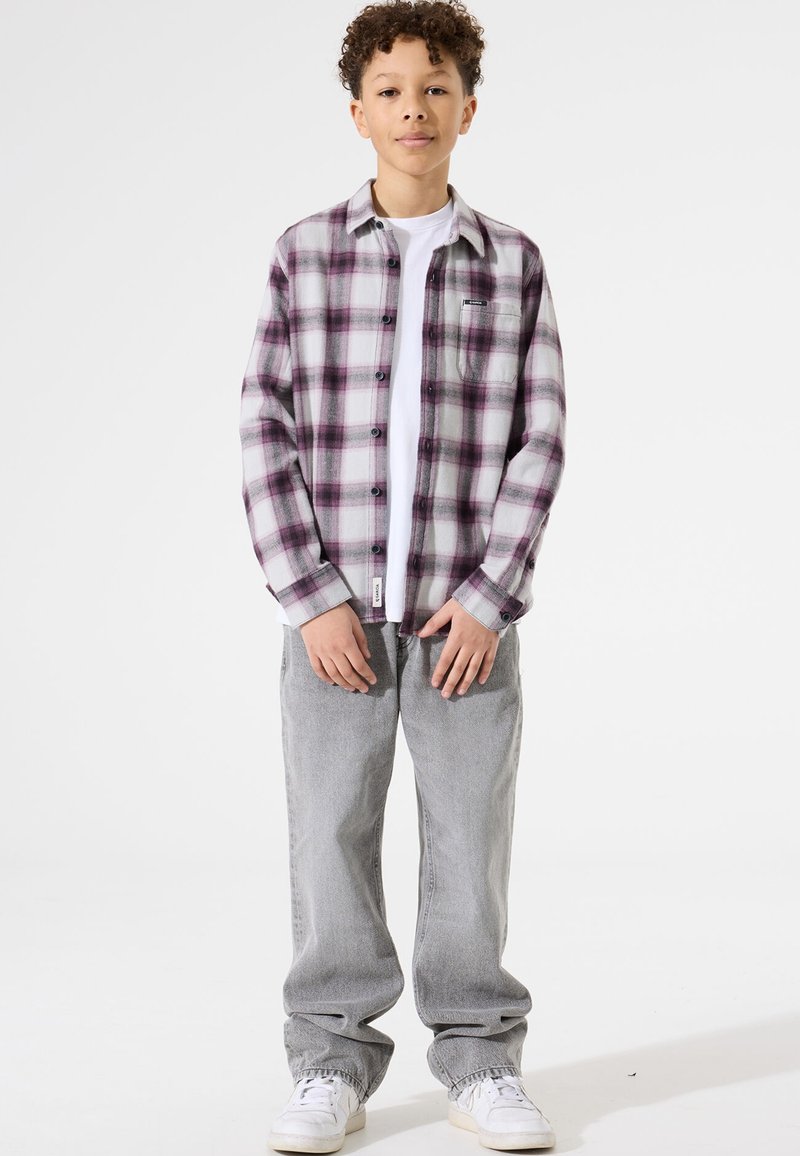Jongen staat gekleed in een paarse en witte geruite overhemden over een wit T-shirt, grijze jeans en witte sneakers, tegen een eenvoudige witte achtergrond.