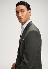 Mężczyzna w ciemnoszarym garniturze i białej koszuli w paski, patrzy w lewo na tle jasnego, gładkiego tła.