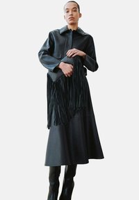 Zwarte leren jas met een kraag, gecombineerd met een lange zwarte rok; een zwarte tas met franjes in de hand. Gladde textuur, minimalistisch ontwerp.