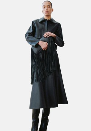 Czarna skórzana kurtka z kołnierzem, zestawiona z długą czarną spódnicą; trzyma frędzlowaną czarną torbę. Gładka faktura, minimalistyczny design.