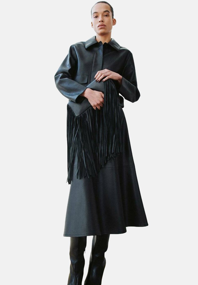 Zwarte leren jas met een kraag, gecombineerd met een lange zwarte rok; een zwarte tas met franjes in de hand. Gladde textuur, minimalistisch ontwerp.