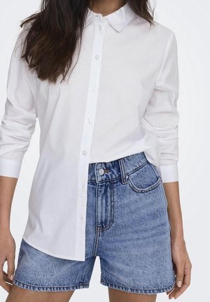 Femme portant une chemise blanche à manches longues boutonnée, rentrée dans un short en denim bleu taille haute, sur un fond uni.