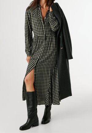 Vrouw draagt een zwart-wit geruite jurk met knopen en een hoge split aan de dij, zwarte kniehoge laarzen, en houdt een zwarte jas over één schouder.
