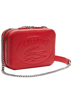 Sac bandoulière rectangulaire en cuir rouge avec logo Lacoste embossé et bandoulière chaîne argentée sur fond blanc.