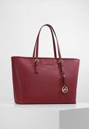 Sac cabas en cuir bordeaux avec doubles poignées et breloque logo Michael Kors dorée suspendue à la poignée droite, sur une surface blanche.