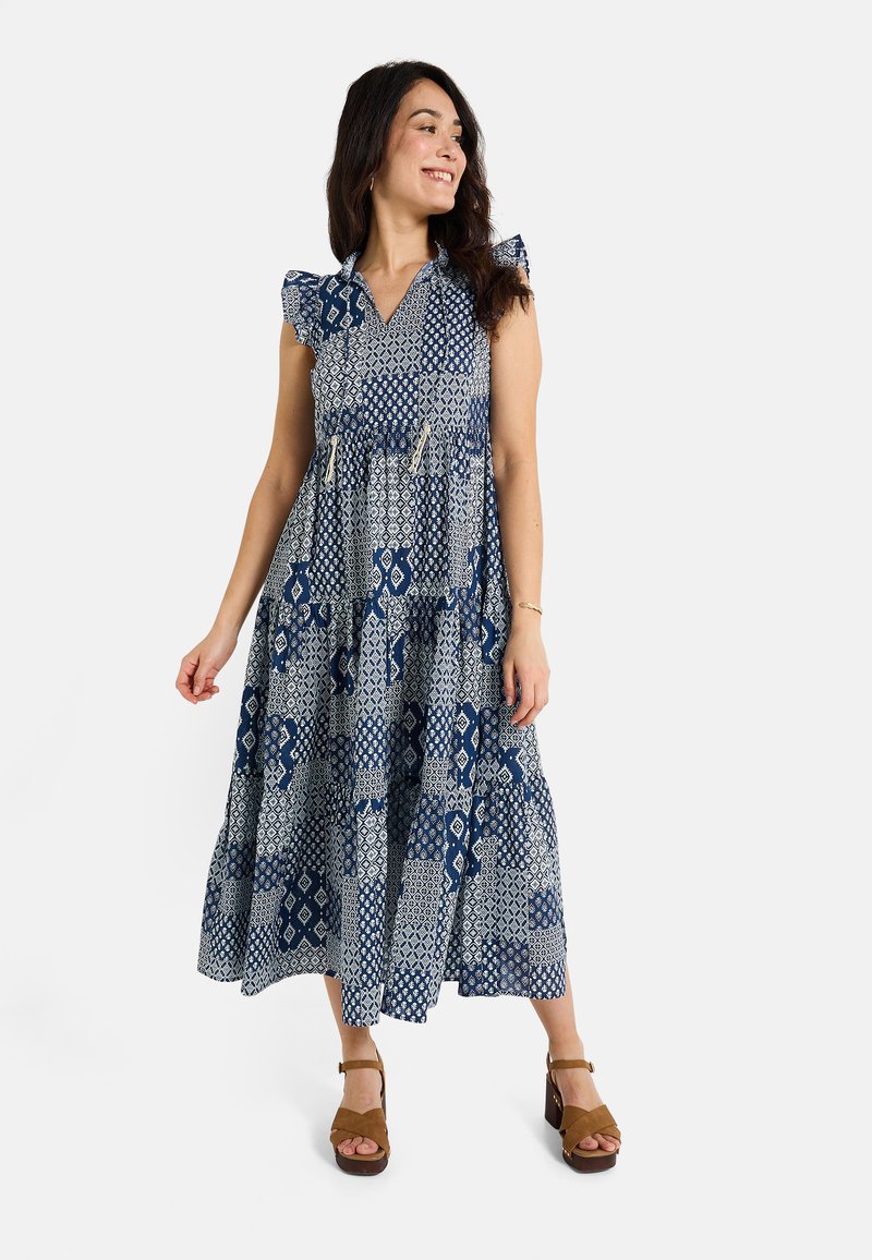 Vestido maxi en azul marino con patrones geométricos blancos, hombros con volantes y diseño en capas. Hecho de material ligero, combinado con tacones marrones.