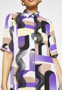 Femme portant une chemise à manches courtes multicolore avec des motifs géométriques abstraits et des coups de pinceau noirs ondulés.