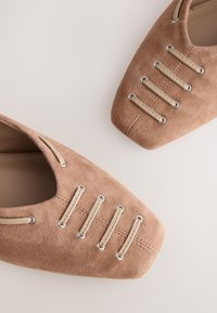 Ballerines en daim marron à bouts carrés avec quatre bandes en cuir beige passées à travers des œillets métalliques sur le dessus.