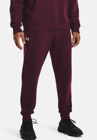 Burgunderfarbene Jogginghosen aus weichem Stoff, mit elastischen Bündchen und dem silbernen Under Armour-Logo am linken Oberschenkel, kombiniert mit schwarzen Sneakers.