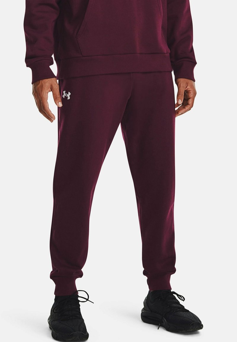 Burgunderfarbene Jogginghosen aus weichem Stoff, mit elastischen Bündchen und dem silbernen Under Armour-Logo am linken Oberschenkel, kombiniert mit schwarzen Sneakers.