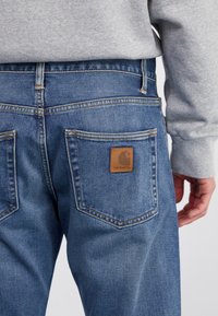 Pantalones vaqueros azules de mezclilla con un acabado texturizado, mostrando dos bolsillos traseros y una etiqueta de cuero marrón con un logo. Combinados con una sudadera gris.