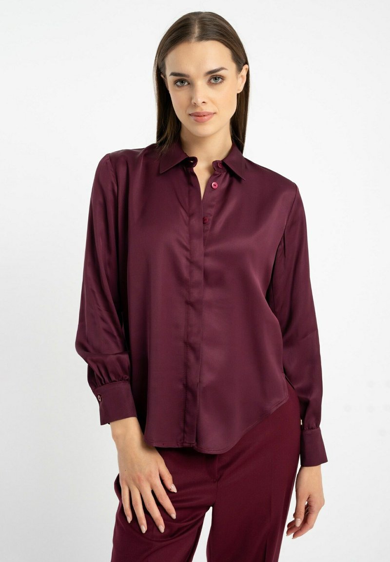 Burgundowa satynowa koszula z wywiniętym kołnierzykiem, guzikami z przodu i długimi rękawami. Gładka tekstura o lekko zaokrąglonym dolnym wykończeniu i mankietami z guzikami.