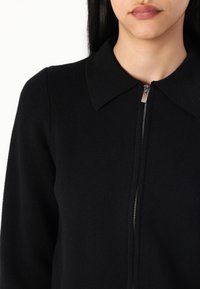 Černý zip-up kousek oblečení s límcem a hladkou texturou, se stříbrným zipem umístěným uprostřed vpředu.