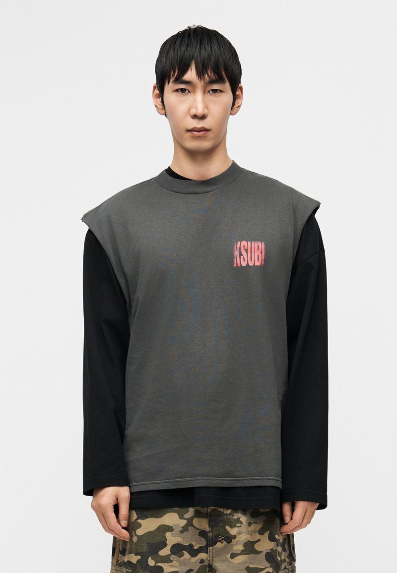 Γκρι φαρδιά πουκάμισο με κοντά μανίκια, με κόκκινο λογότυπο στο αριστερό στήθος· φορεμένο πάνω από μαύρο πουκάμισο με μακριά μανίκια. Σορτς καμουφλάζ.