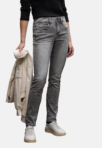 Jeans de denim gris con un corte ajustado, cinco bolsillos y un leve desgaste. Combinados con una chaqueta bomber de color beige claro y utilizados con zapatillas blancas.